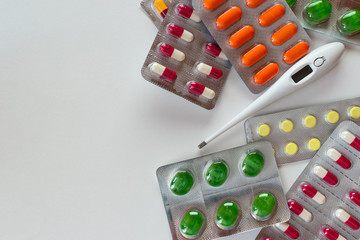 Blisters with multi-colored capsules and pills, electronic thermometer on a white table.
