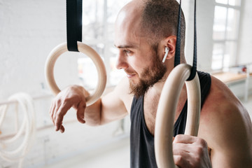 Male bearded athlete with modern headphones resting and holding gymnastics rings at light gym. Fit...