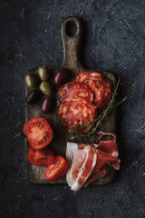 Appetizers on cutting board. Chorizo, jamon, olives and tomato on dark background top view.