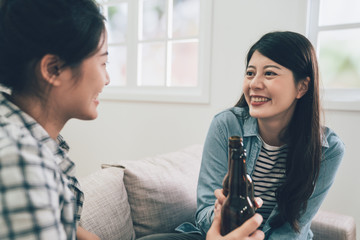 Fototapeta premium two asian female friends drinking beers together sitting on comfort couch sofa in living room in dormitory. girl roommates looking each other cheers making toast together celebrating new relationship
