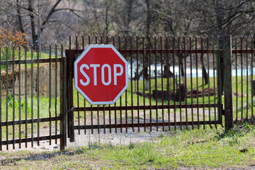 Partially rusted metal locked picket fence driveway doors with large stop road sign mounted in front surrounded with uncut grass and tall trees in background on warm sunny spring day