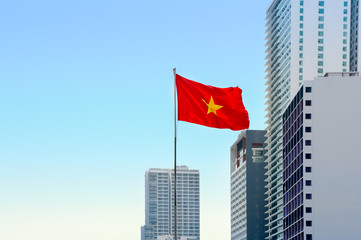 The national flag of Vietnam on a high spire swaying in the wind. Blue sky and skyscrapers on background. 