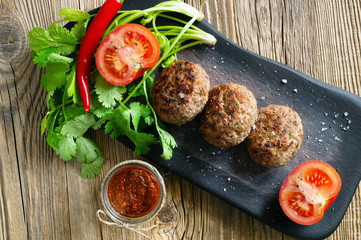 fried meat patties, tomato halves, hot pepper, and cilantro on black serving board with adjika sauce in a glass jar on a wooden background