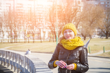 Outdoor close up portrait of blond curly playful hipster woman on street, looking at camera, smiling at city parks. Model wearing white sweater, yelow winter hat, sarf, gloves.