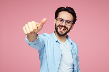 Portrait of trendy young bearded man in blue stylish shirt and eyeglasses showing thumb up over pink background