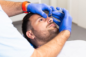 Handsome young man is getting a rejuvenating facial injections. He is sitting calmly at clinic. The expert beautician is filling male wrinkles by hyaluronic acid.