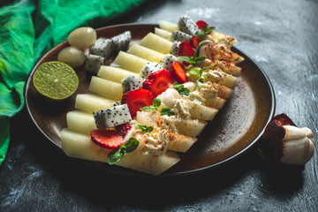 on a dark, stone table lies a plate with a prepared meal of exotic fruits: honey melon, rambutan, dragon fruit, mangosteen with the addition of chilli peppers, strawberries and whipped cream