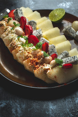 on a dark, stone table lies a plate with a prepared meal of exotic fruits: honey melon, rambutan, dragon fruit, mangosteen with the addition of chilli peppers, strawberries and whipped cream