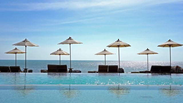 Tropical resort scene, infinity pool and umbrellas over beach chairs looking over calm ocean waters