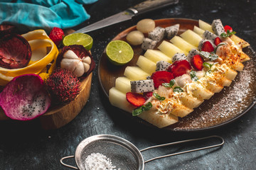 on a dark, stone table lies a plate with a prepared meal of exotic fruits: honey melon, rambutan, dragon fruit, mangosteen with the addition of chilli peppers, strawberries and whipped cream, sprinkle