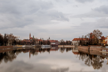 Fototapeta na wymiar Calm dreamy view of buildings, trees, and gray sky reflected in a crystal clear water of Odra river in Wroclaw, Poland. Typical buildings with red roofs on a background