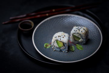 avocado and cream cheese inside out maki sushi wuth sesame seeds on dark plate with chopsticks and soy sauce