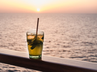 Beautiful glass, bright cocktail with ice and straw, standing on the open deck against the backdrop of sea waves and sunset. Side view, close-up. Concept of leisure and travel