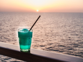 Beautiful glass, bright cocktail with ice and straw, standing on the open deck against the backdrop of sea waves and sunset. Side view, close-up. Concept of leisure and travel