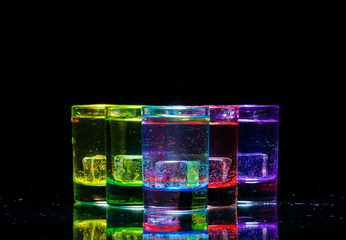 Multicolored glasses filled with alcoholic drinks, with ice cubes inside, standing on the mirror surface. Black background. Conceptual, celebrated design