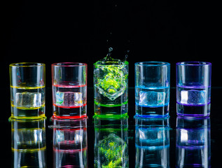 Multicolored glasses filled with alcoholic drinks, with splases of ice cubes falling inside, standing on the mirror surface. Black background. Conceptual, celebrated, commercial design