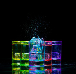 Multicolored glasses filled with alcoholic drinks, with splases of ice cubes falling inside, standing on the mirror surface. Black background. Conceptual, celebrated, commercial design