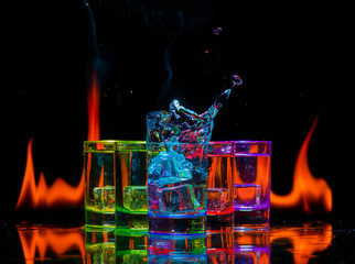 Multicolored glasses filled with alcoholic drinks, with splases of ice cubes falling inside, standing on the mirror surface on background of fire flames. On black. Conceptual design