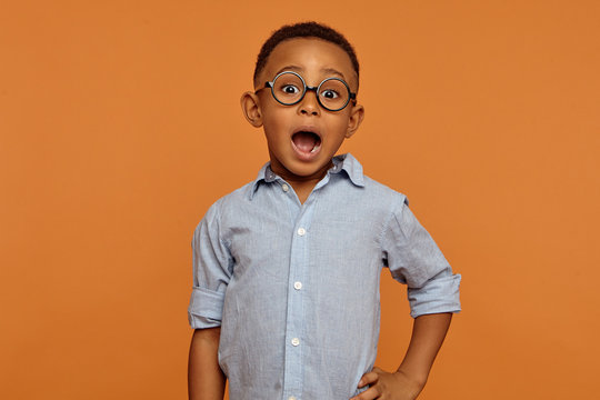 Isolated Picture Of Handsome Emotional Bug Eyed Dark Skinned Schoolboy Wearing Stylish Glasses Expressing Shock, Keeping Mouth Wide Opened. Excitement, Fascination And True Genuine Emotions