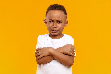 Portrait of stubborn reluctant black dark skinny child posing in studio with grumpy dissatisfied grimace, keeping arms on his chest, demonstrating unwillingness to clean his room. Body language