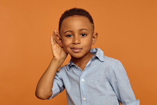 Overhear, spy and secrecy concept. Isolated studio portrait of cute nosy African ten year old boy spying on someone, keeping hand at his ear and listening attentively, trying to hear everything