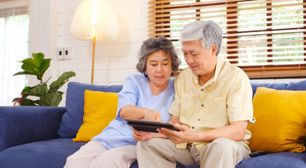 Happy senior asian couple using digital tablet computer sitting on sofa at home living room background, senior people and technology lifestyles