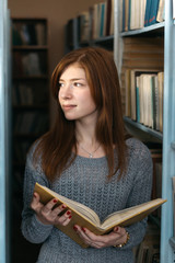 The girl with the red hair in the library stands with a book in her hands between the bookshelves and looks at the light.