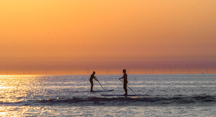 Two men silhouette approaching to each other on paddle surfing at sunset on the Zandvoort beach in the Netherlands  - obrazy, fototapety, plakaty