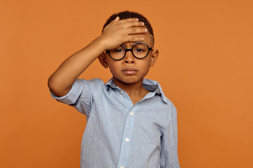 Sad unhappy African American schoolboy in trendy eyewear having painful look, holding hand on his forehead after he fell. Frustrated sick dark skinned child suffering form terrible headache