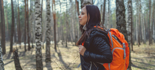 Fototapeta na wymiar Traveling woman with backpack in woods Side view of brunette standing with bright orange backpack in tranquil sunny forest looking away