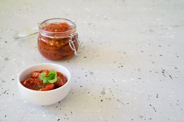 Sun-dried tomatoes in olive oil a glass jar and a white bowl with basil on a gray background with copy space