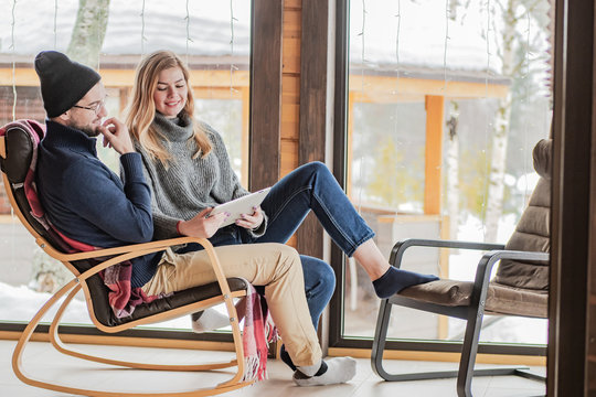 A Married Couple Are Sitting In A Chair. A Man Shows On The Tablet News To His Girlfriend. Rest On The Veranda In Winter. Family Couple On The Background Of The Window. Living Outside The City.