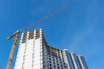 high-rise construction crane with a long arrow of yellow color against the blue sky over a new multi-storey building of concrete and brick under construction