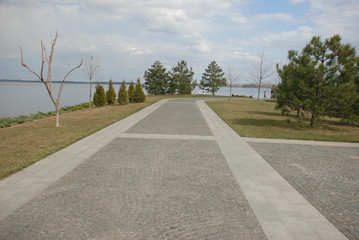 A gray granite walkway runs in a park along the river. Around grow trees and shrubs.