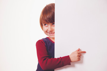 Funny little child girl laughing behind a white board and pointing with her finger towards copy space area. Space for text.