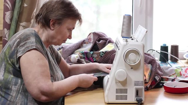 Mature woman sews patchwork quilt on sewing machine at home. Side view.