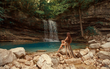Fototapeta na wymiar mysterious river nymph on bare rocks near small blue lake in Martvili Canyon Lagoon and wonderful waterfall, girl with red hair in long brown dress with long train cut. no model face, creative colors
