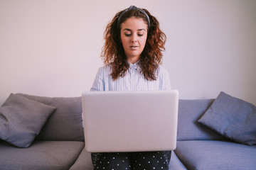 A businesswoman is sitting on a sofa with her laptop on her legs. Look at the computer screen while writing something. She has dark hair and wears a ribbon in her hair. 