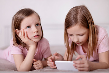 Two little girls (sisters) in pink pajamas on the bed watching a smartphone and reading an e-book.