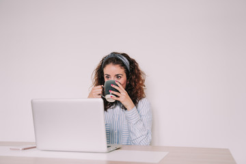 A young woman is sitting in front of her computer while drinking a cup of coffee. Look at something pretentiously on the screen of your laptop. 