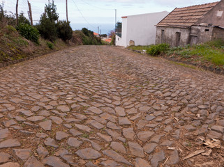 Eine Straße aus Kopfsteinpflaster führt durch ein halb verlassenes Dorf im Westen von Madeira. Im Hintergrund kann man den Atlantischen Ozean sehen.