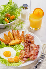 English breakfast-plate with fried bacon, with French fries, fried egg stands on a white table and a glass of fresh juice.
