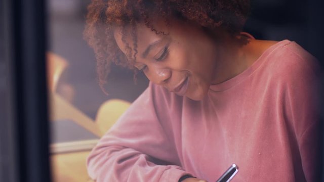 Close up of happy young African woman writing letter indoor by the window. Joyful  black young woman smiling and writing on the book. Young people study education lifestyle 4k clip