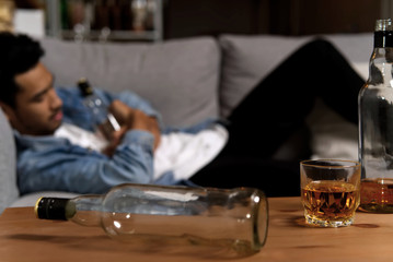Drunk man sleeping on sofa and holding bottle of whiskey after he drank a lot of alcohol in one night. Some of bottle and glass put on table showing how much he had drunk. Alcoholism concept.