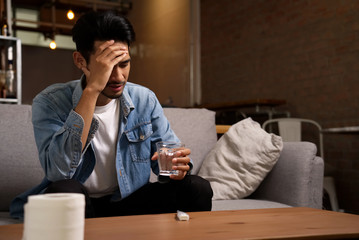 Illness and unhealthy condition concept. Asian man sitting on sofa having headache holding glass of water and putting his hand on the head. He has high body temperature after checking by thermometer.