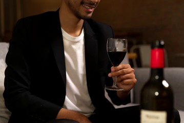 Luxury alcohol drinking. Crop shot of man sitting on sofa holding glass of wine with smile before drinking. Bottle of wine put on table as foreground. Alcohol drinking and party concept.
