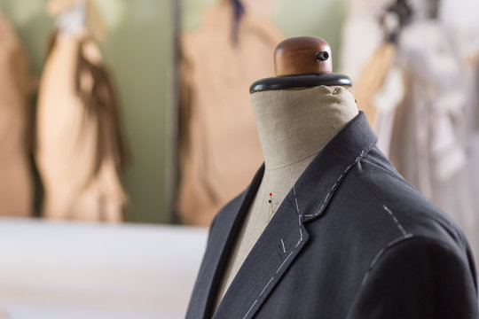 Tailored Suit Jacket With Alterations Marked With Basting Stitch On A Mannequin At Bespoke Tailor's Workshop. Variety Of Patterns Hanging In The Background.