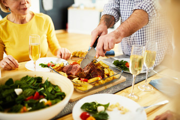 Close-up of unrecognizable man in checkered shirt standing at table and cutting cooked meat for dinner during dinner party with friends