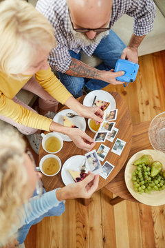 Directly above view of mature friends sitting on sofa and talking while watching polaroid images made at party, they drinking tea and eating cake