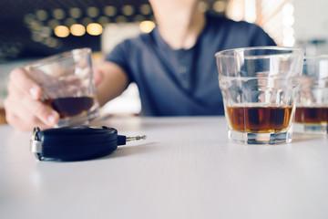 Close up on car keys on the table with a man holding a glass of drink in the background drinking and driving